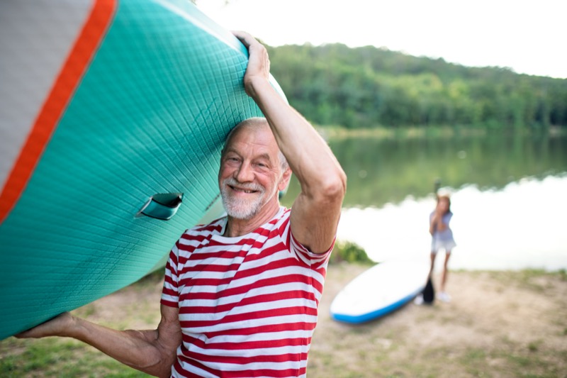 Senior man carrying paddleboard by lake in summer, looking at camera.